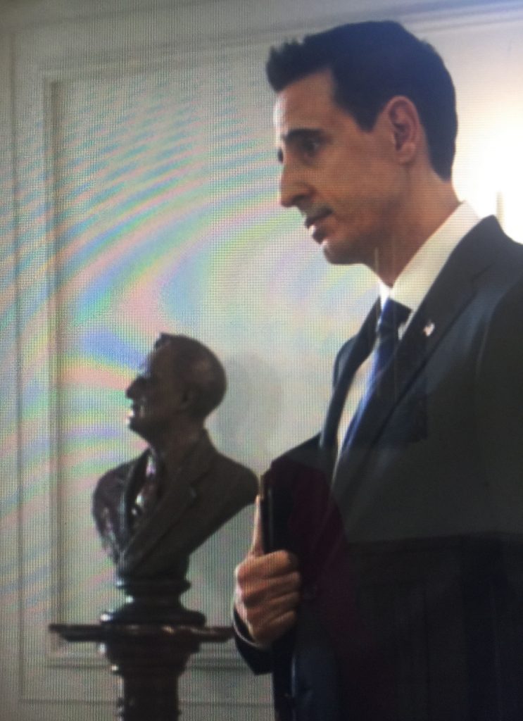 A man in a suit and tie standing next to a bust.